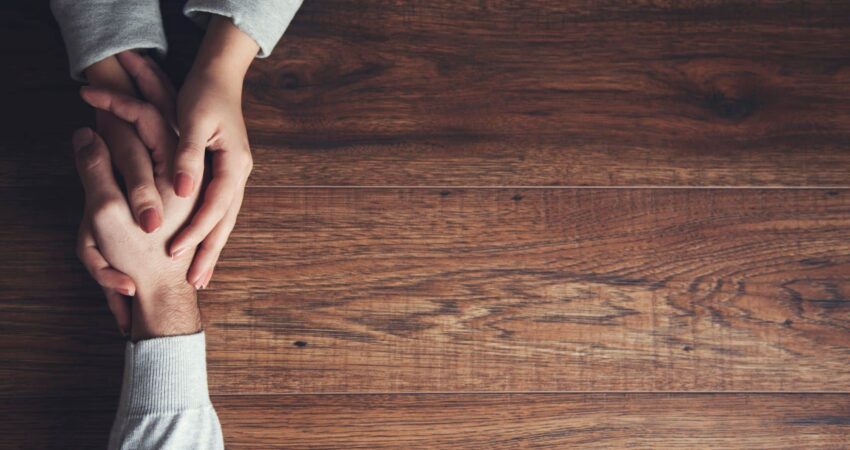 Ruskea pöytä, jonka päällä kahden ihmisen kädet pitelevät toisistaan kiinni.