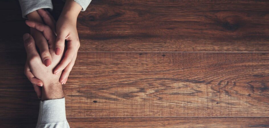 Ruskea pöytä, jonka päällä kahden ihmisen kädet pitelevät toisistaan kiinni.