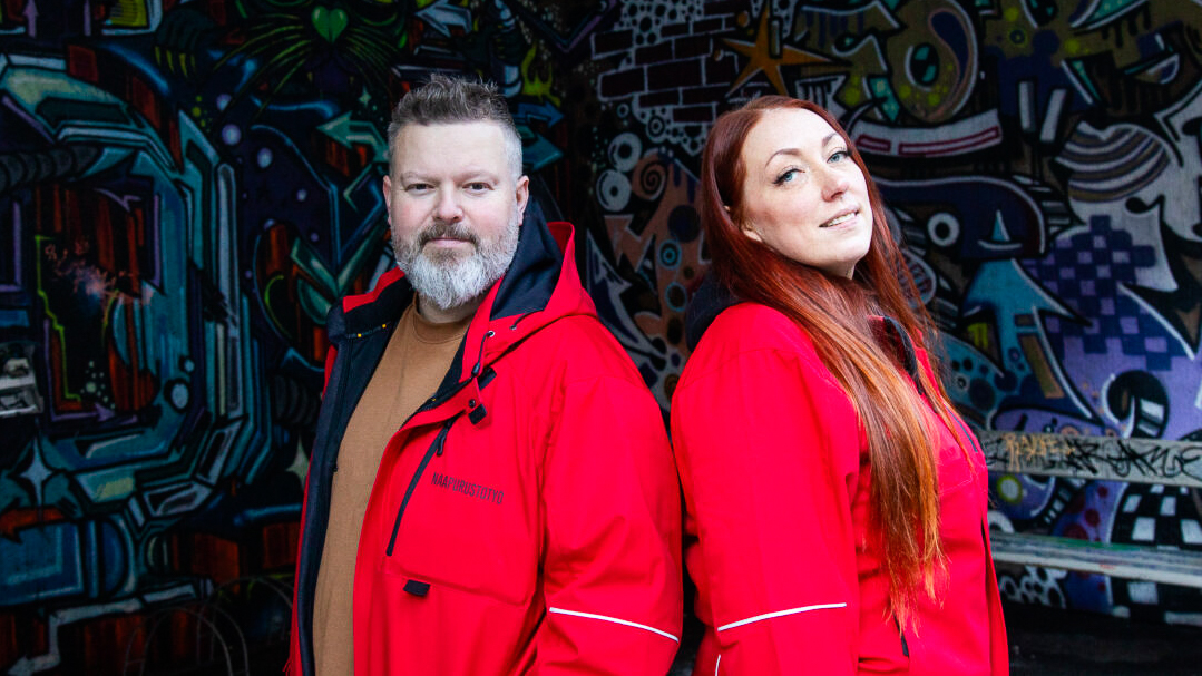 Kaksi henkilöä seisoo graffitiseinän edessä ja katsoo kameraan. heillä on kirkkaanpunaiset takit