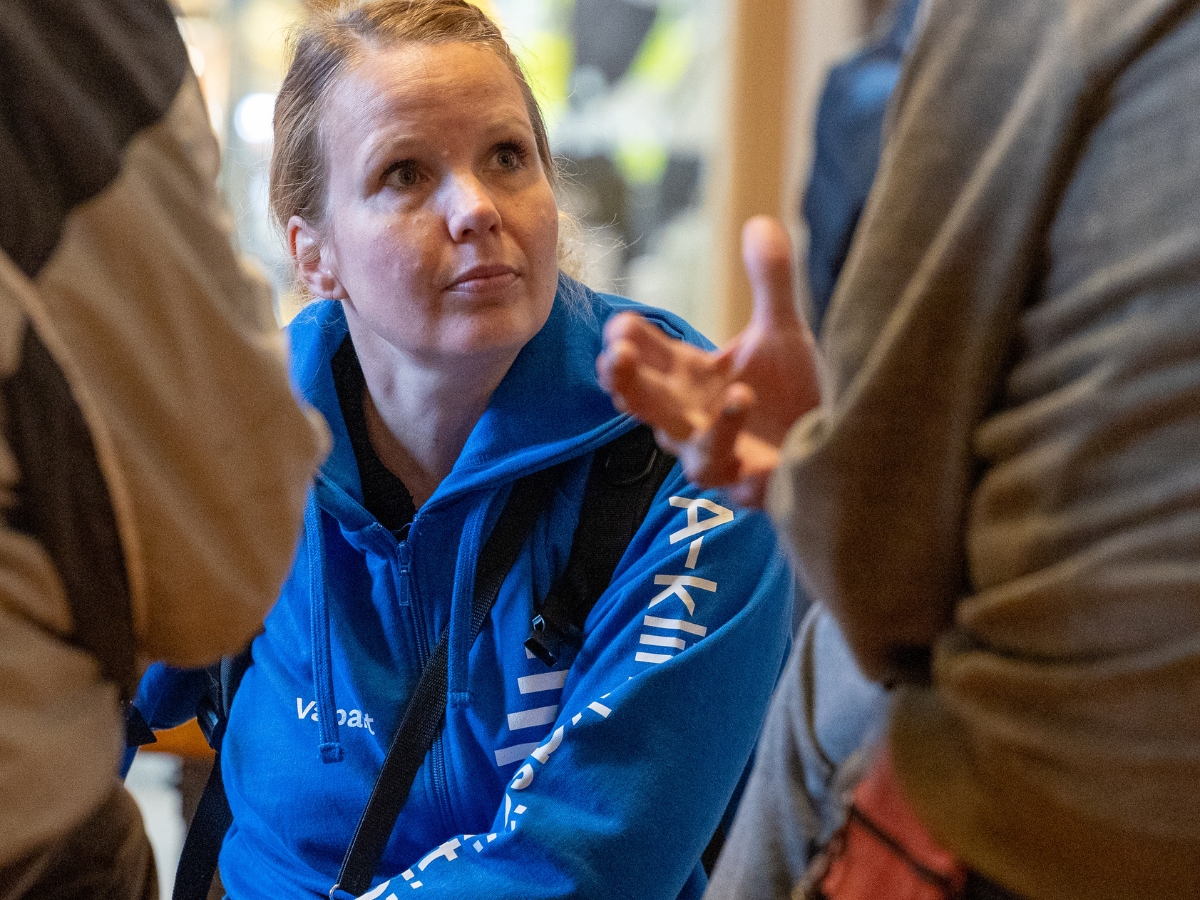 Henkilö kuuntelee keskittyneesti keskustelua sisätiloissa. Hänellä on yllään sininen huppari, jossa lukee "Vaparit", ja kaulassaan avainnauha. Etualalla näkyy osittain kaksi henkilöä, joista toinen elehtii käsillään puhuessaan.