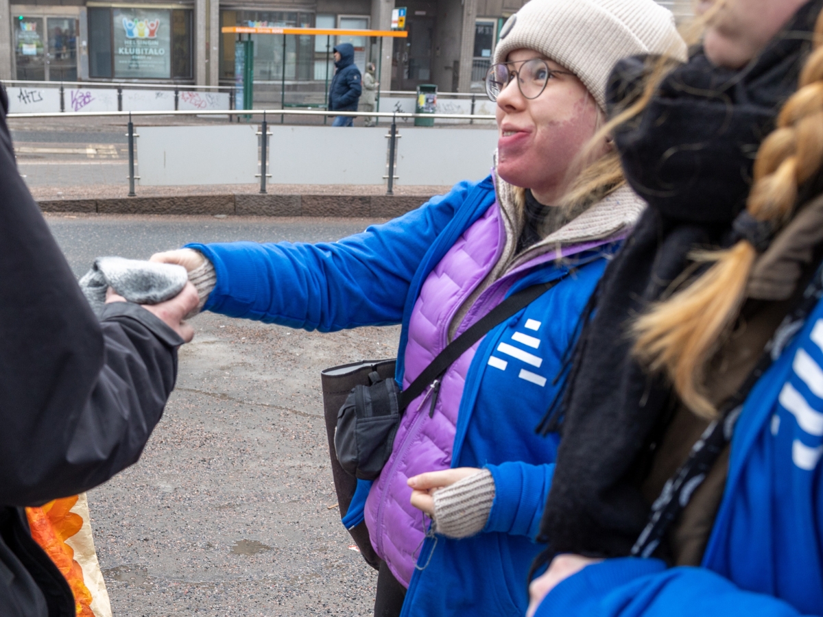 Henkilö, jolla on valkoinen pipo, silmälasit ja sininen takki, ojentaa harmaita käsineitä toiselle henkilölle ulkona kadulla. Hänellä on myös violetti toppaliivi ja olkalaukku. Vieressä seisoo toinen henkilö sinisessä takissa ja mustassa huivissa. Taustalla näkyy kaupungin katuympäristö, pysäkki ja ohikulkijoita.