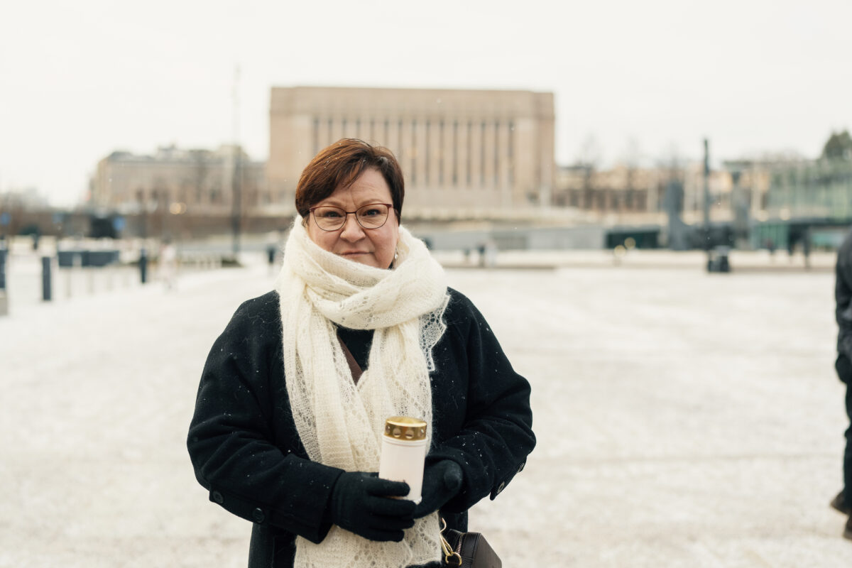 Kuvassa nainen valkoisessa kaulahuvissa seisoo aukealla ja katsoo kameraan. Hänellä on kädessään palava hautakynttilä