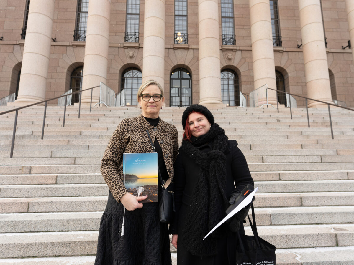 Kuvassa kaksi henkilöä seisoo Eduskuntatalon portailla ja pitelee suruadressia