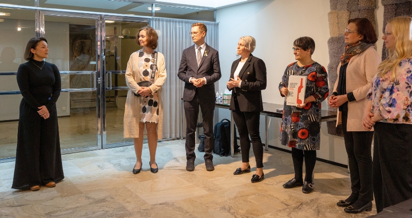 Kuvassa Näkymättömät lapset -vetoomuksen luovutus eduskuntaryhmien edustajille Eduskunnassa. Kansanedustajia seisoo puolikaaressa kuuntelemassa kokemustoimijan puhetta