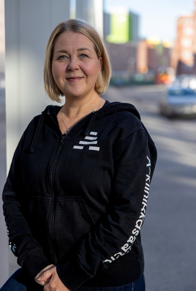 Kuvassa vaaleahiuksinen henkilö seisoo kadun reunassa ja katsoo kameraan. Hänellä on yllään musta huppari, jossa lukee A-klinikkasäätiö