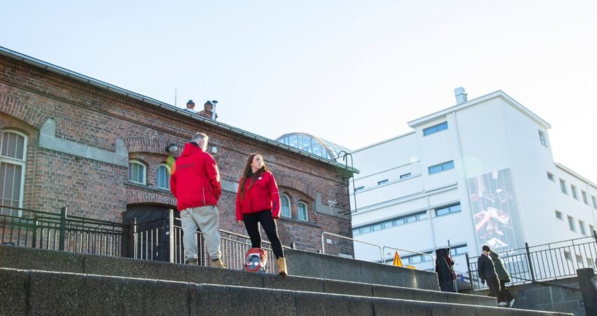 Kuvassa kaksi henkilöä seisoo portailla ja katsoo eri suuntiin. Heillä on päällään kirkkaanpunaiset takit