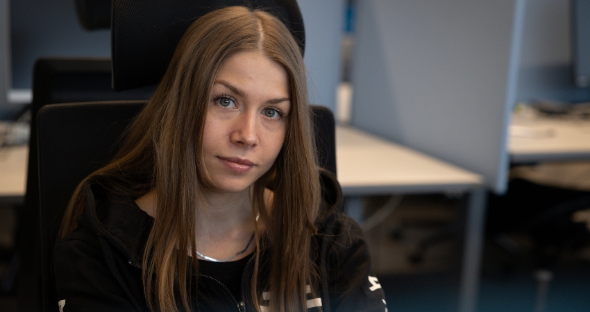 Kuvassa pitkähiuksinen henkilö katsoo suoraan kameraan toimistoympäristössä