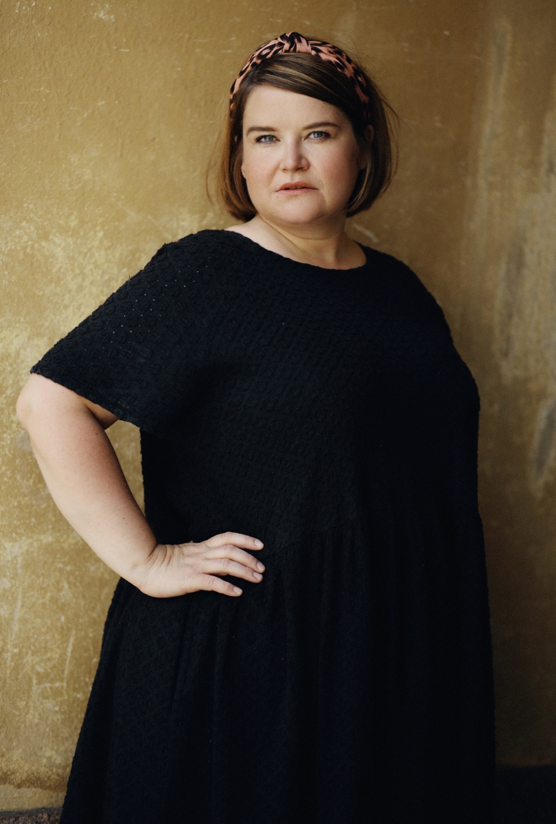 Nainen seisoo nojaamassa seinää vasten. Hänellä on yllään musta, lyhythihainen mekko ja päässään leopardikuosinen hiuspanta. Hän katsoo suoraan kameraan vakavalla ilmeellä. Taustalla on karkearakenteinen, lämminsävyinen seinä.