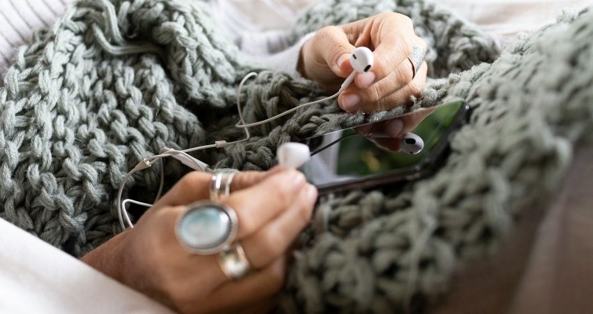 Henkilö istuu pehmeän, vihreän neulepeiton alla ja pitää käsissään valkoisia nappikuulokkeita sekä älypuhelinta. Tilanne huokuu rauhallisuutta ja hetkeä ennen meditaation tai äänitteen kuuntelua.