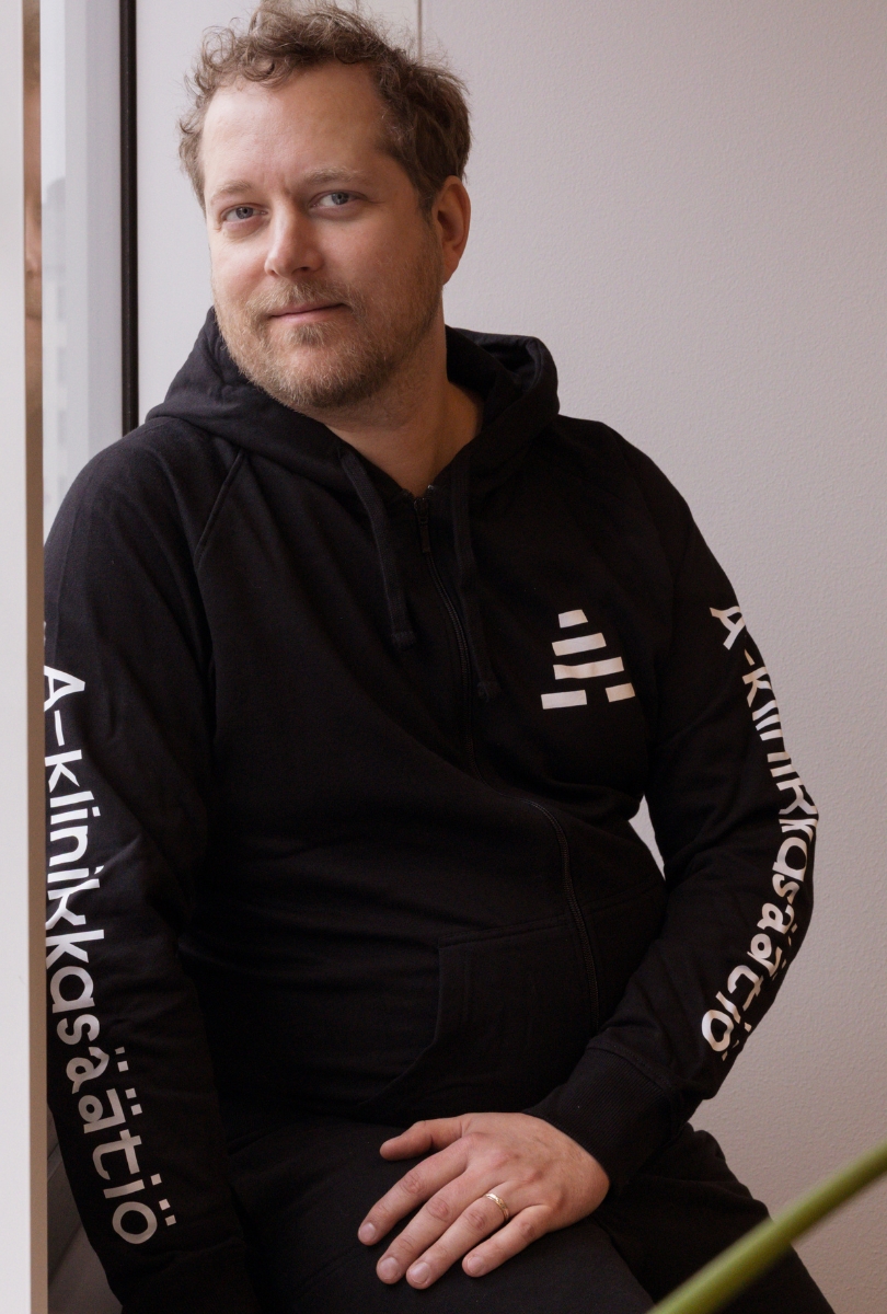 Henkilö istuu ikkunan vieressä ja katsoo kameraan rauhallisesti hymyillen. Hänellä on yllään musta huppari, jonka hihoissa ja rinnassa lukee A-klinikkasäätiö-logoteksti ja -tunnus.