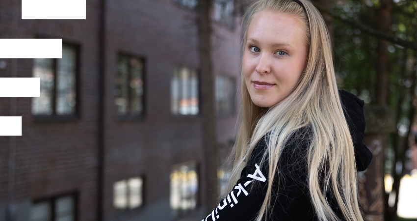 Henkilö katsoo kameraan ulkona kaupunkimaisessa ympäristössä. Hänellä on pitkät vaaleat hiukset ja päällään musta huppari, jossa lukee A-Klinikkasäätiö. Taustalla näkyy tiilirakennus ja puita.