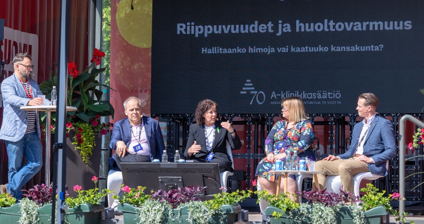 Paneelikeskustelu käynnissä SuomiAreena-tapahtumassa ulkolavalla kesäisessä säässä. Vasemmalla juontaja seisoo pöydän ääressä, oikealla neljä paneelikeskustelijaa istuu lavalla. Taustalla suuri näyttö, jossa lukee "Riippuvuudet ja huoltovarmuus – Hallitaanko himoja vai kaatuuko kansakunta?" sekä A-klinikkasäätiön 70-vuotisjuhlalogo. Lavalla kukkaistutuksia ja yleisölle suunnattuja istumapaikkoja. Tilanne vaikuttaa aktiiviselta ja keskustelevalta.