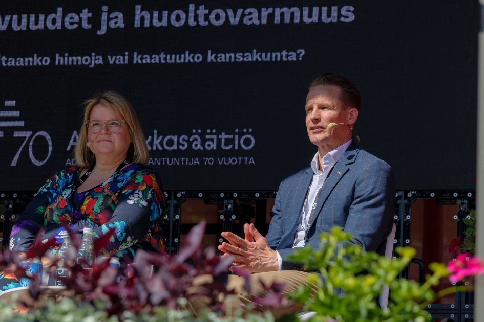 Kaksi henkilöä istuu lavalla SuomiAreena-tapahtumassa keskustelemassa auringonpaisteessa. Taustalla näkyy suuri näyttö, jossa lukee "Riippuvuudet ja huoltovarmuus – Hallitaanko himoja vai kaatuuko kansakunta?" sekä A-klinikkasäätiön 70-vuotisjuhlalogo. Vasemmalla oleva henkilö kuuntelee rauhallisesti ja on pukeutunut värikkääseen kukkakuosiseen asuun. Oikealla oleva henkilö puhuu ja elehtii käsillään, yllään sininen ruudullinen bleiseri ja valkoinen kauluspaita. Etualalla näkyy istutuksia ja kukkia.
