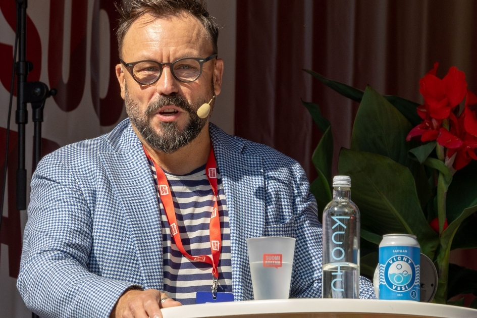 Henkilö puhuu lavalla SuomiAreena-tapahtumassa ja käyttää puhelinkuulokemikrofonia. Hänellä on yllään sinivalkoinen ruudullinen bleiseri ja raidallinen paita sekä punainen tapahtumapassi kaulassa. Henkilö seisoo korkean pöydän ääressä, jonka päällä on vesipullo, virvoitusjuoma ja tapahtumalogolla varustettu muovimuki. Taustalla näkyy punaisia kukkia ja tapahtuman somisteita. Aurinko valaisee henkilön kasvot kirkkaasti.