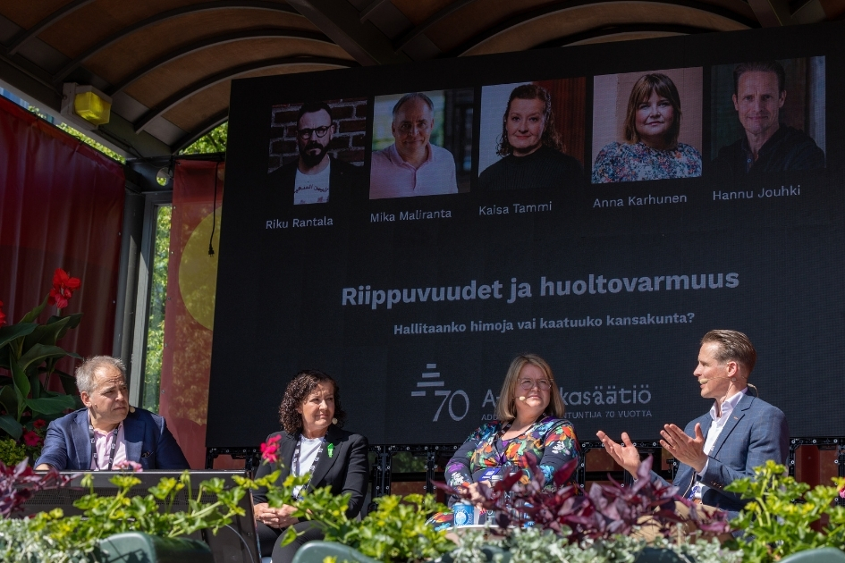 Paneelikeskustelu käynnissä ulkolavalla SuomiAreena-tapahtumassa. Neljä henkilöä istuu rivissä lavalla, taustanaan suuri näyttö. Oikeanpuoleisin henkilö puhuu ja elehtii käsillään, muut kuuntelevat keskittyneesti. Näytöllä näkyy keskustelun otsikko "Riippuvuudet ja huoltovarmuus – Hallitaanko himoja vai kaatuuko kansakunta?", A-klinikkasäätiön 70-vuotisjuhlalogo sekä viiden keskusteluun osallistuvan henkilön nimet ja kasvokuvat: Riku Rantala, Mika Maliranta, Kaisa Tammi, Anna Karhunen ja Hannu Jouhki. Etualalla on värikkäitä kukkia ja istutuksia.