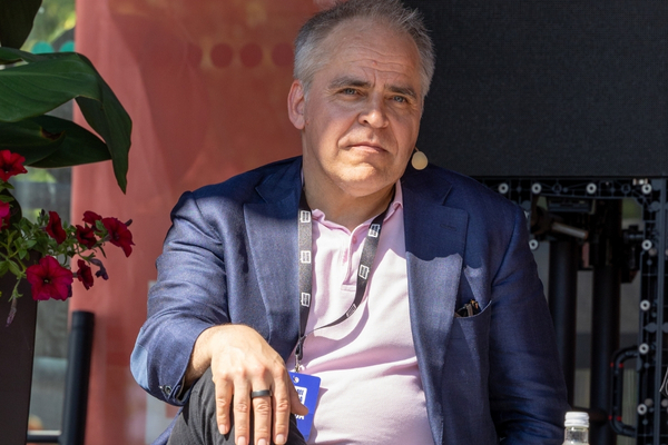 Henkilö istuu lavalla SuomiAreena-tapahtumassa ja katsoo vakavana kameraan. Hänellä on yllään sininen pikkutakki ja vaaleanpunainen kauluspaita, ja kaulassaan tapahtuman tunniste. Mies käyttää puhelinkuulokemikrofonia ja hänen vierellään on punaisia kukkia. Taustalla näkyy osittain musta näyttö ja tapahtumatekniikkaa. Aurinko paistaa kirkkaasti kasvoille.
