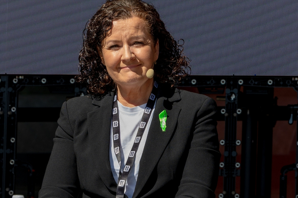 istuu lavalla SuomiAreena-tapahtumassa ja hymyilee rauhallisesti. Hänellä on tummat, kiharat hiukset ja yllään musta jakku sekä vaalea paita. Kaulassa näkyy tapahtuman tunniste ja rintapielessä vihreä nauha. Nainen käyttää puhelinkuulokemikrofonia. Taustalla näkyy osa näyttölaitteistosta ja teknistä kalustoa. Aurinko valaisee hänen kasvonsa kirkkaasti.