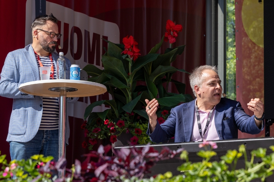Kaksi henkilöä osallistuu keskusteluun SuomiAreena-tapahtuman lavalla ulkotilassa. Vasemmalla seisova henkilö on pukeutunut raitapaitaan ja vaaleansiniseen bleiseriin ja nojaa korkeaan pöytään, jonka päällä on juomapulloja. Oikealla istuva henkilö elehtii käsillään puhuessaan ja käyttää puhelinkuulokemikrofonia; hänellä on yllään sininen pikkutakki ja vaaleanpunainen paita. Taustalla näkyy punaisia kukkia ja suuri kasvi koristeena sekä osa tapahtuman visuaalisesta somistuksesta. Etualalla kukkaistutuksia.