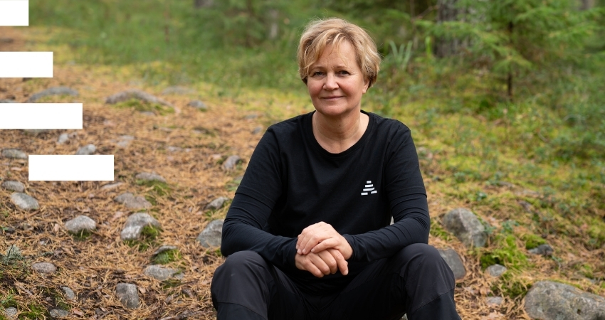 Vaaleahiuksinen henkilö istuu metsäpolulla, katsoo kameraan ja nojaa käsillä polviinsa. Hänellä on yllään A-klinikkasäätiön musta huppari ja kuvan vasemmassa reunassa on graafinen elementti, joka on puolikas valkea A-tunnus