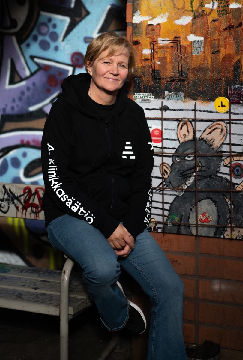 Vaaleahiuksinen henkilö istuu korkealla jakkaralla graffitiseinän edessä ja katsoo kameraan pieni hymy huulillaan. Hänellä on yllään A-klinikkasäätiön musta huppari.