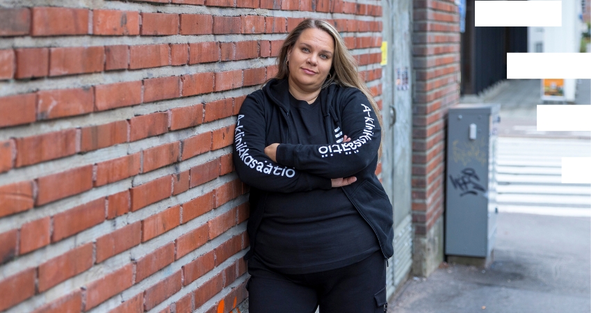 seisoo käsivarret ristissä punatiiliseinää vasten kaupunkikadulla. Hänellä on yllään musta huppari, jossa lukee A-klinikkosäätiö, sekä mustat housut. Taustalla näkyy sähkökaappi ja suojatie.