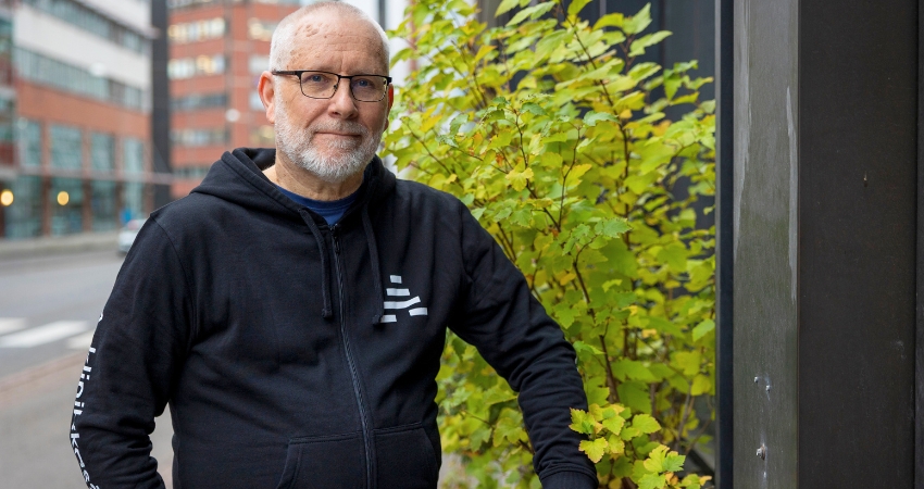 Silmälasipäinen henkilö seisoo kadulla seinän lähellä ja katsoo kameraan. Hänellä on A-klinikkasäätiön musta huppari yllään