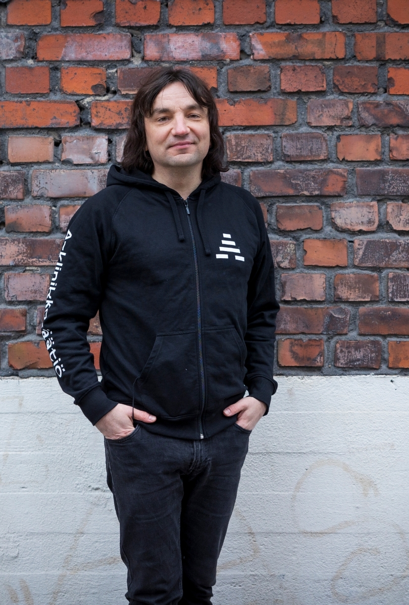 Kuvassa tummahiuksinen henkilö katsoo kameraan ja hänellä on yllään A-klinikkasäätiön musta huppari. Taustalla on tiiliseinä