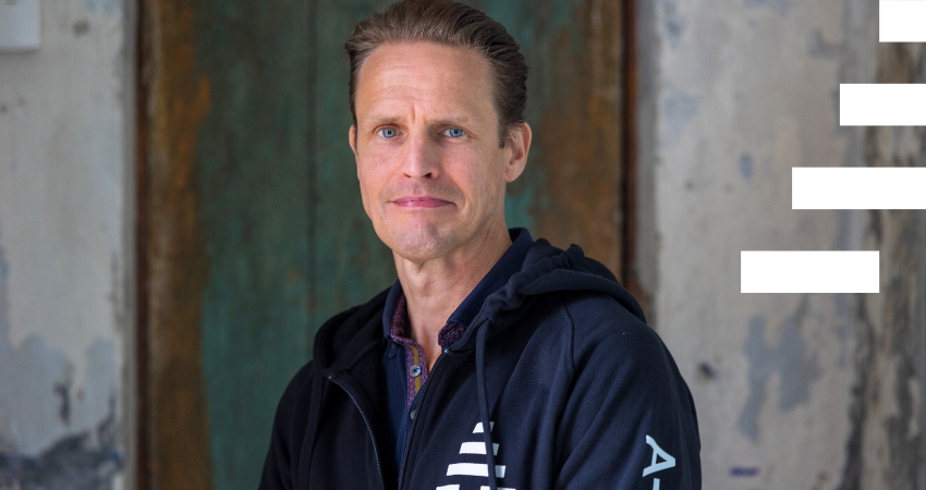 Henkilö katsoo kameraan ja istuu vanhan oven edessä. Hänellä on yllään A-klinikkasäätiön musta huppari