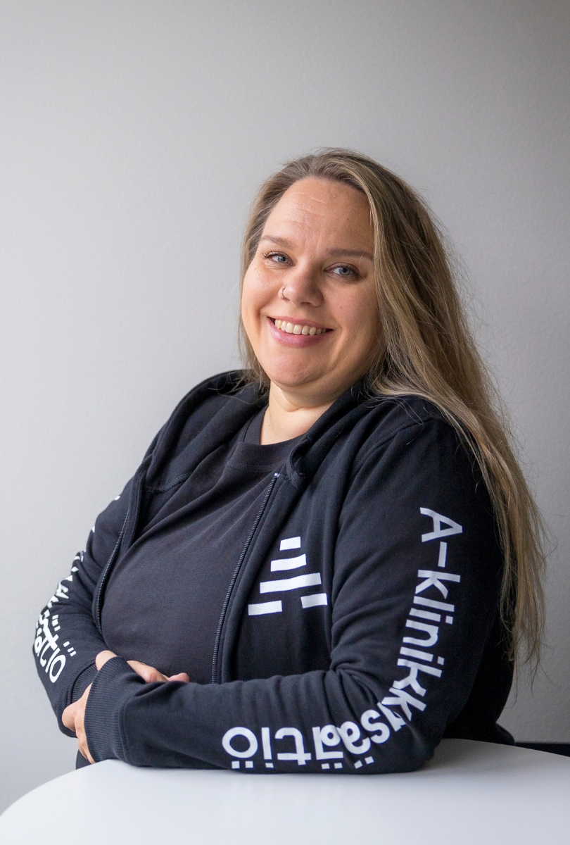 Kuvassa pitkähiuksinen henkilö nojaa pöytään ja katsoo kameraan hymyillen. Hänellä on yllään musta A-klinikkasäätiön huppari