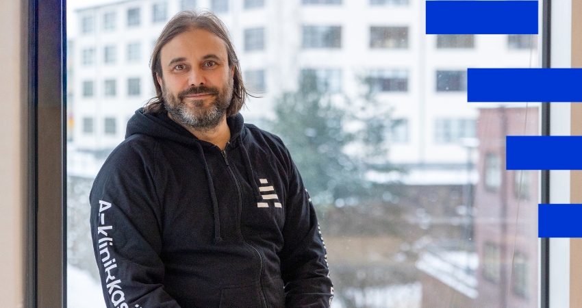 Henkilö istuu ikkunalaudalla ja katsoo kameraan. Hänellä on tummat hiukset ja parta sekä yllään A-klinikkasäätiön musta huppari. Kuvan oikeassa laidassa on graafisena elementtinä puolikas A-klinikkasäätiön sininen A-tunnus