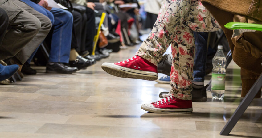 Paljon ihmisiä odotushuoneessa. Kuvattu lattiatasosta, jolloin kuvassa näkyy vain odottavien ihmisten kenkiä ja jalkoja