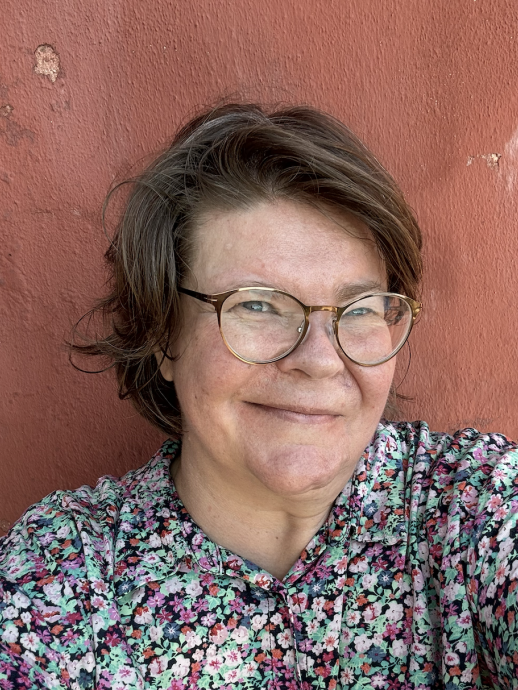 Kuvassa silmälasipäinen henkilö kukkapaidassa katsoo kameraan. Hän seisoo tiilenpunaisen seinän edessä