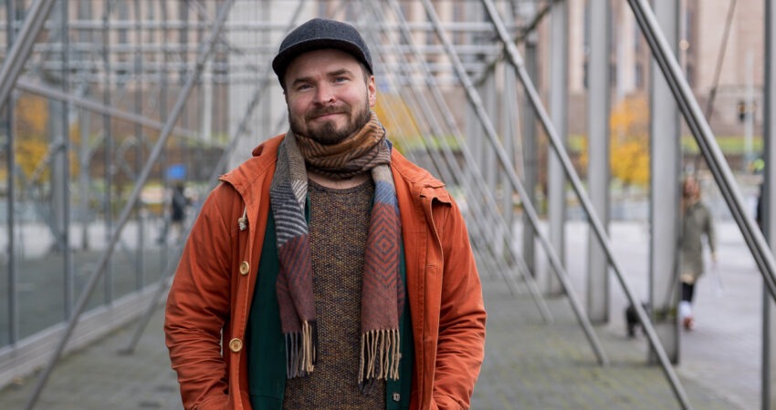 Kuvassa henkilö, jolla on oranssi takki ja kaulahuivi katsoo kameraan Sanomatalon pylväiden luona. Hänellä on hattu päässään ja kuva on otettu ulkona. Taustalla näkyy eduskuntatalo