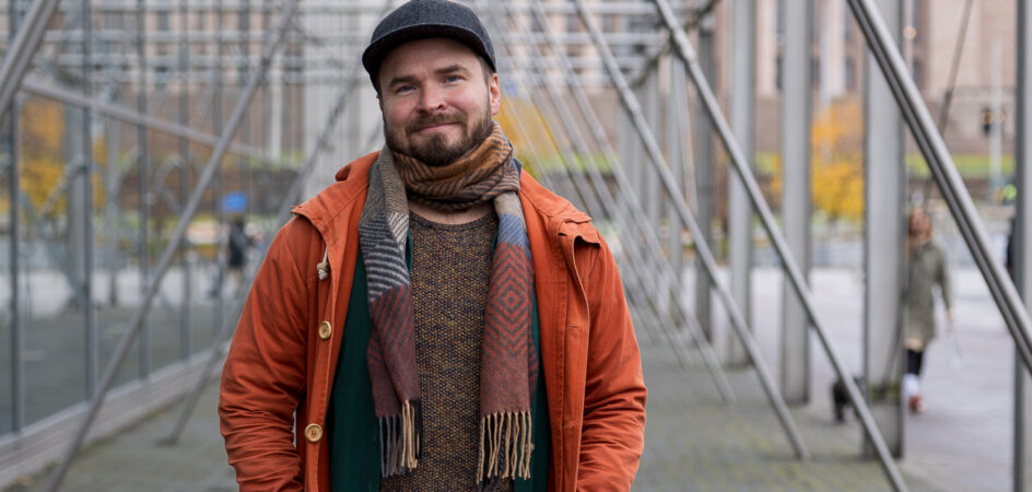 Kuvassa henkilö, jolla on oranssi takki ja kaulahuivi katsoo kameraan Sanomatalon pylväiden luona. Hänellä on hattu päässään ja kuva on otettu ulkona. Taustalla näkyy eduskuntatalo