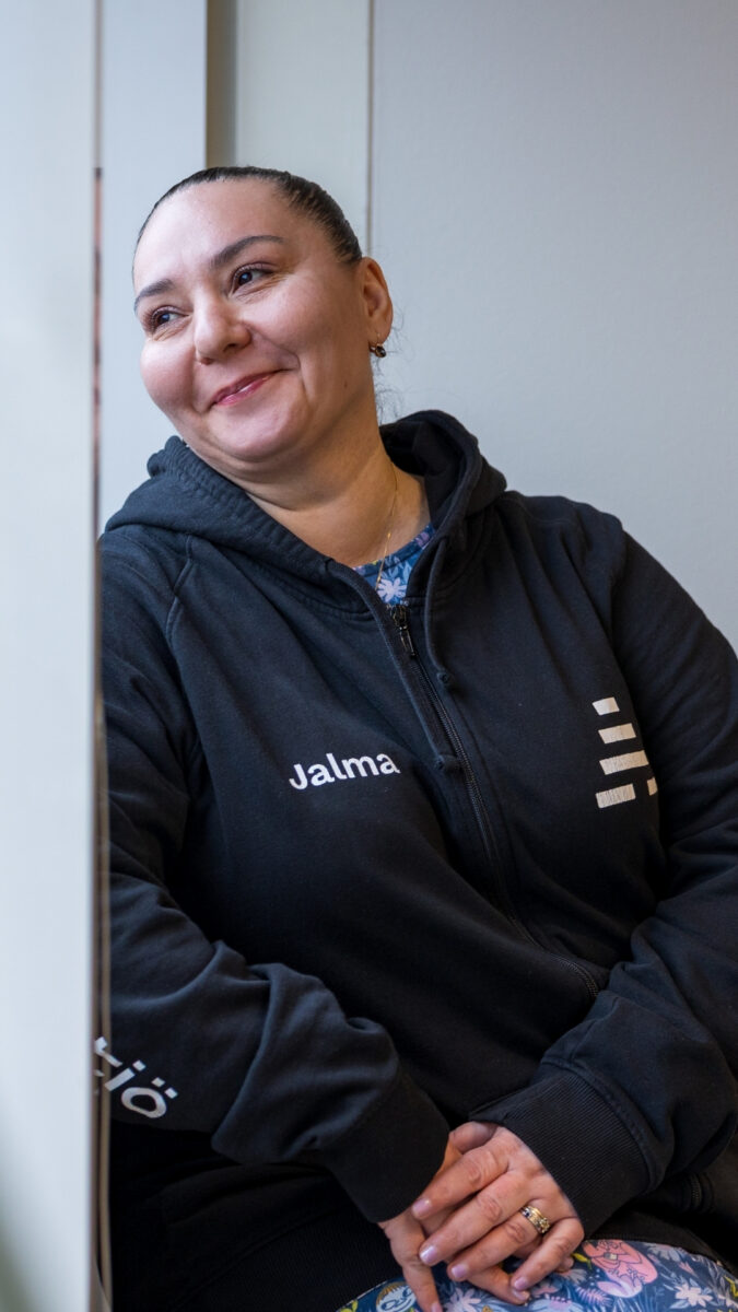 Kuvassa henkilö istuu ikkunalaudalla ja katsoo hymyillen ulos. Hänellä on yllään musta huppari, jossa lukee edessä Jalma ja hihoissa A-klinikkasäätiö