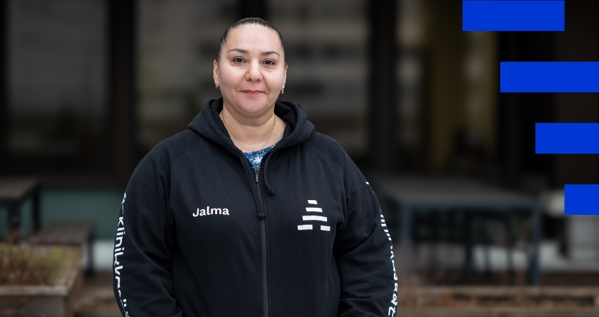 Kuvassa henkilö seisoo ulkona talon edessä ja katsoo kameraan hymyillen. Hänellä on yllään musta huppari, jossa edessä lukee Jalma ja hihoissa A-klinikkasäätiö