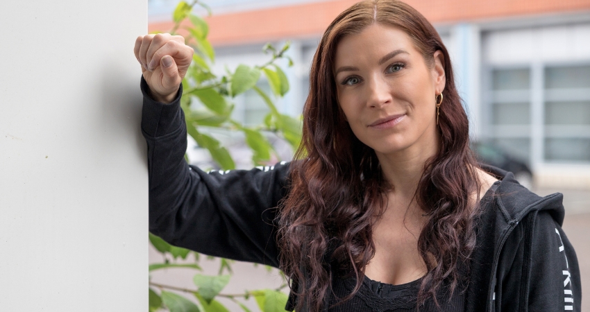 Kuvassa henkilö, jolla on pitkät ruskeat hiukset nojaa ulkona pylvääseen toisella kädellä ja katsoo hymyillen kameraan. Hänellä on yllään musta huppari, jossa lukee A-klinikkasäätiö. Taustalla on tiiliseinää ja vihreitä lehtiä