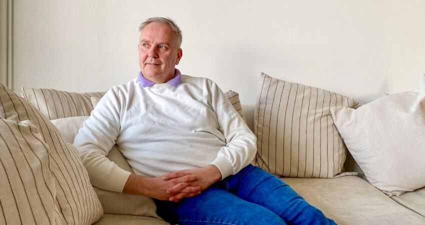 Kuvassa henkilö istuu sohvalla ja katsoo vasemmalle ikkunasta ulos. Hänellä on yllään vaalea paita sekä siniset farkut. Sohva on vaalea ja siinä on raidallisia koristetyynyjä