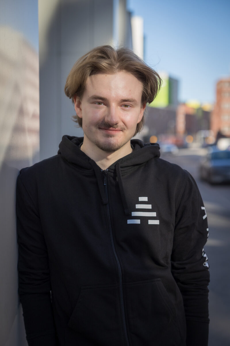 Kuvassa vaaleahiuksinen henkilö katumaisemassa. Hän katsoo kameraan ja hymyilee. Henkilöllä on yllään musta huppari, jossa lukee A-klinikkasäätiö