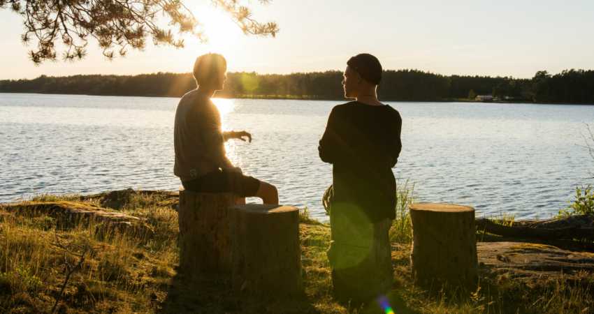 kuvassa kaksi henkilöä keskustelee järven rannalla ilta-auringossa. heidät on kuvattu vasten aurinkoa, joten näkyy vain silhuetit