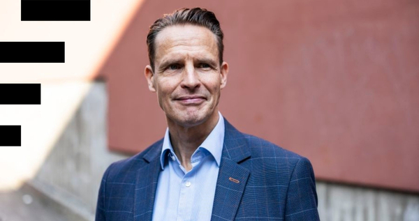 Kuvassa henkilö sinisävyisessä puvussa katsoo oikealle yläviistoon. Taustalla on tiilenpunainen rapattu seinä