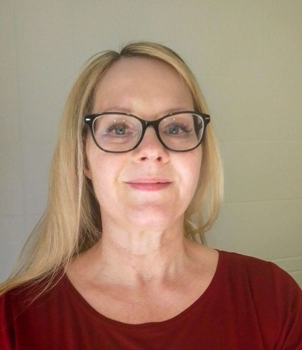 Kuvassa vaaleahiuksisen naisen kasvokuva. Hänellä on silmälasit ja punainen paita