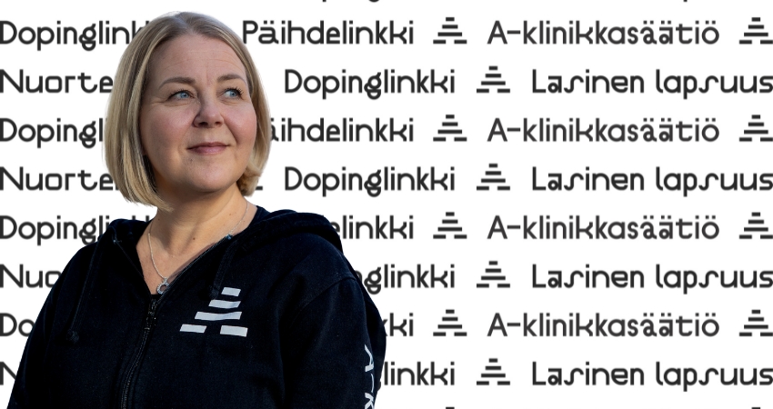 Kuvassa A-klinikkasäätiön asiantuntija seisoo A-klinikkasäätiön logotapetin edessä. Taustalla näkyy Lasinen lapsuus, Dopinglinkki, Nuortenlinkki ja Päihdelinkki