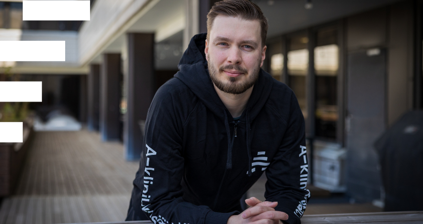 kuvassa henkilö nojaa ulkona pöytään ja katsoo kameraan. hänellä on lempeä hymy, tummat hiukset, parta ja yllään A-klinikkasäätiön huppari