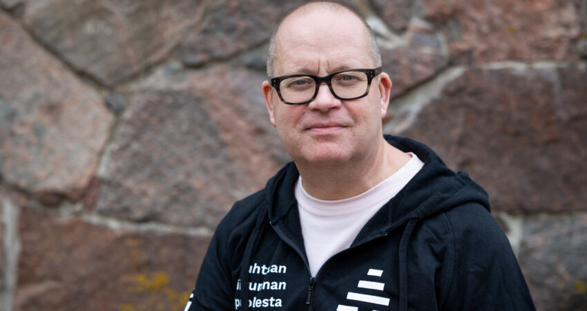 Henkilö seisoo ulkona kiviseinän edessä, katsoo kameraan ja hymyilee kevyesti. Hänellä on mustat silmälasit ja musta huppari, jossa on A-klinikkasäätiön logo sekä valkoista tekstiä.