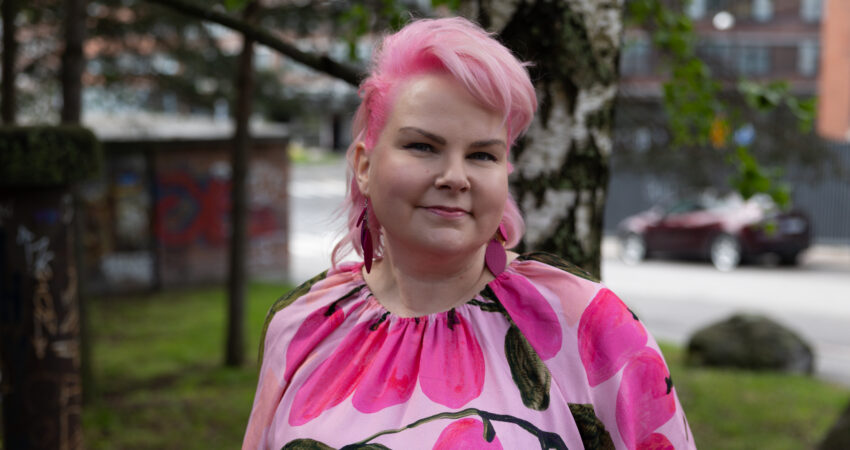 Tanni Karvonen katsoo ystävällisellä ilmeellä kameraan. Tannilla on vaaleanpunaiset hiukset sekä yllään vaaleanpunainen mekko ja korvikset.