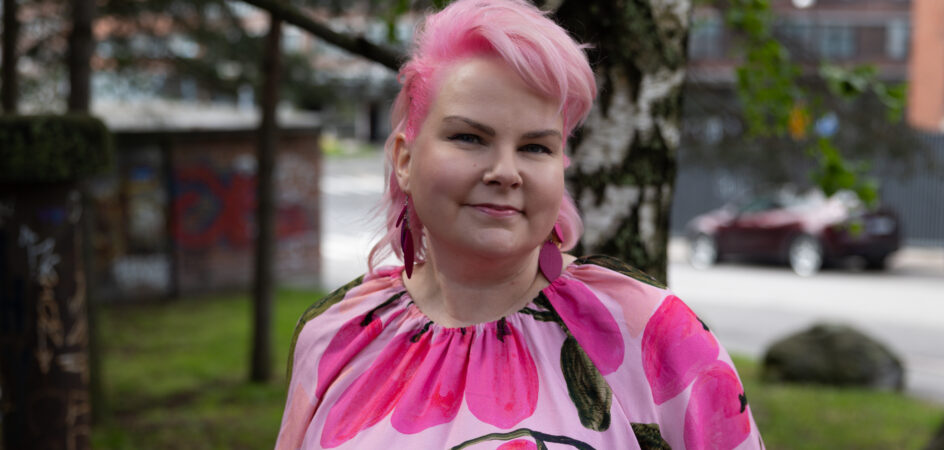 Tanni Karvonen katsoo ystävällisellä ilmeellä kameraan. Tannilla on vaaleanpunaiset hiukset sekä yllään vaaleanpunainen mekko ja korvikset.